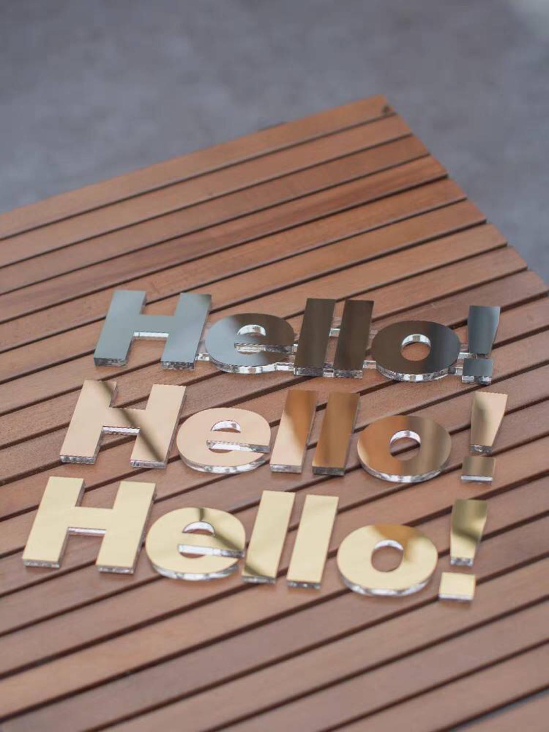 Hello! signage three lines in yellow gold, rose gold, and black metal acrylic finihs with clear acrylic backing laying on a wooden table
